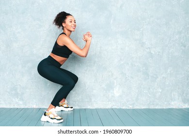 Fitness smiling black woman in sports clothing with afro curls hairstyle.She doing squats. Young beautiful model with perfect tanned body.Female in studio near gray wall