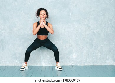 Fitness smiling black woman in sports clothing with afro curls hairstyle.She doing squats. Young beautiful model with perfect tanned body.Female in studio near gray wall. Happy and cheerful  - Powered by Shutterstock