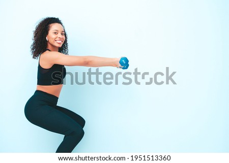 Fitness smiling black woman in sports clothing with afro curls hairstyle.She doing squats. Young beautiful model with perfect tanned body.Female holding dumbbells in studio near light blue wall