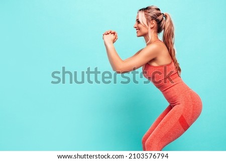 Fitness smiling woman in pink sports clothing. Young beautiful model with perfect body.Female posing near blue wall in studio.Cheerful and happy. Stretching out before training. Doing squats  