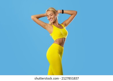 Fitness And Sport Concept. Portrait Of Smiling Young Woman Wearing Bracelet Tracker And Sportswear Posing Isolated On Blue Studio, Enjoying Her Perfect Slim Body Raising Arms Holding Hands Behind Head - Powered by Shutterstock