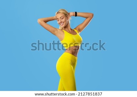 Fitness And Sport Concept. Portrait Of Smiling Young Woman Wearing Bracelet Tracker And Sportswear Posing Isolated On Blue Studio, Enjoying Her Perfect Slim Body Raising Arms Holding Hands Behind Head