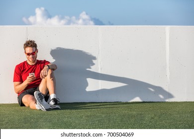 Fitness Sport Man Using Mobile Phone App After Workout Training Relaxing Sitting On Outdoor Grass Post Run. Smartwatch Tech Device Online App Concept. Active And Health Person.