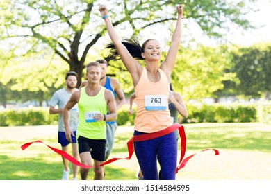 Fitness, Sport And Success Concept - Happy Woman Winning Race And Coming First To Finish Red Ribbon Over Group Of Sportsmen Running Marathon With Badge Numbers At Park