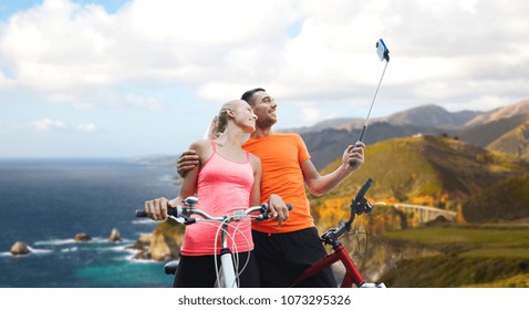 Fitness, Sport And Technology Concept - Happy Couple With Bicycle Taking Picture By Smartphone On Selfie Stick Over Big Sur Hills And Pacific Ocean Background In California