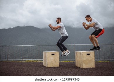 fitness, sport, training, exercising and people concept - two male friends doing extreme box jumps over misty mountains and sky background. - Powered by Shutterstock