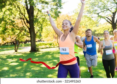 Fitness, Sport, Victory, Success And Healthy Lifestyle Concept - Happy Woman Winning Race And Coming First To Finish Red Ribbon Over Group Of Sportsmen Running Marathon With Badge Numbers Outdoors