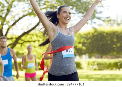 Fitness, Sport, Victory, Success And Healthy Lifestyle Concept - Happy Woman Winning Race And Coming First To Finish Red Ribbon Over Group Of Sportsmen Running Marathon With Badge Numbers Outdoors