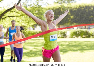 fitness, sport, victory, success and healthy lifestyle concept - happy woman winning race and coming first to finish red ribbon over group of sportsmen running marathon with badge numbers outdoors