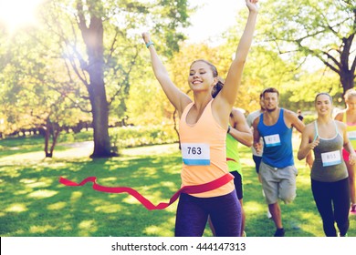 Fitness, Sport, Victory, Success And Healthy Lifestyle Concept - Happy Woman Winning Race And Coming First To Finish Red Ribbon Over Group Of Sportsmen Running Marathon With Badge Numbers Outdoors