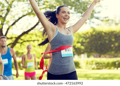 Fitness, Sport, Victory, Success And Healthy Lifestyle Concept - Happy Woman Winning Race And Coming First To Finish Red Ribbon Over Group Of Sportsmen Running Marathon With Badge Numbers Outdoors