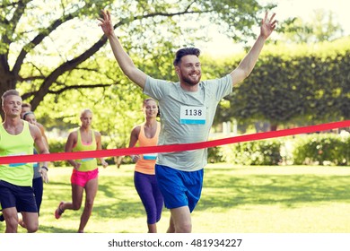 Fitness, Sport, Victory, Success And Healthy Lifestyle Concept - Happy Man Winning Race And Coming First To Finish Red Ribbon Over Group Of Sportsmen Running Marathon With Badge Numbers Outdoors