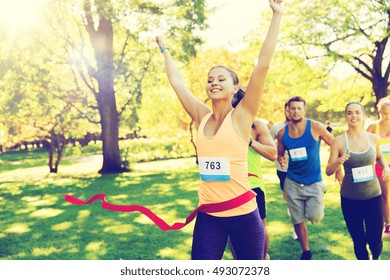 Fitness, Sport, Victory, Success And Healthy Lifestyle Concept - Happy Woman Winning Race And Coming First To Finish Red Ribbon Over Group Of Sportsmen Running Marathon With Badge Numbers Outdoors