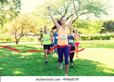 Fitness, Sport, Victory, Success And Healthy Lifestyle Concept - Happy Woman Winning Race And Coming First To Finish Red Ribbon Over Group Of Sportsmen Running Marathon With Badge Numbers Outdoors