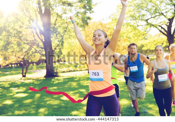 fitness, sport, victory, success and healthy\
lifestyle concept - happy woman winning race and coming first to\
finish red ribbon over group of sportsmen running marathon with\
badge numbers outdoors