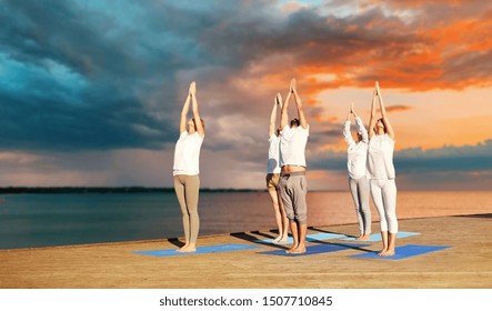 fitness, deporte, yoga y el concepto de estilo de vida saludable - grupo de personas haciendo saludo ascendente posan sobre el muelle del mar sobre el fondo de la puesta del sol