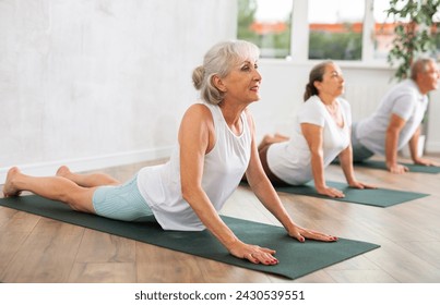 In fitness studio with small group of friends, elderly woman performs asana snake - cobra - pose dog muzzle up, Urdhva Mukha Shvanasana- Bhujangasana. Mature slender woman participates in yoga classes - Powered by Shutterstock