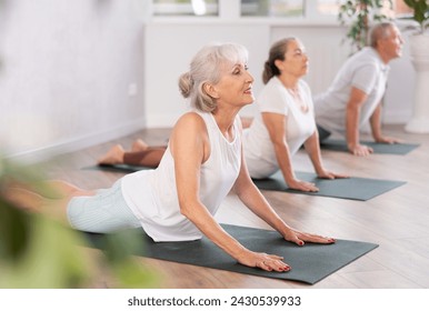 In fitness studio with small group of friends, elderly woman performs asana snake - cobra - pose dog muzzle up, Urdhva Mukha Shvanasana- Bhujangasana. Mature slender woman participates in yoga classes - Powered by Shutterstock