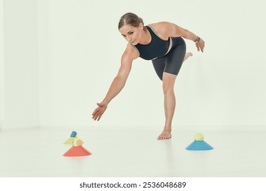 A fitness trainer demonstrates a balance and coordination exercise, reaching out towards cones and tennis balls. Ideal for concepts related to fitness, stability, and body control.. - Powered by Shutterstock