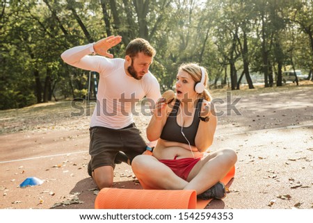 A fitness trainer is getting ready to hit a woman exerciser who is eating junk food.