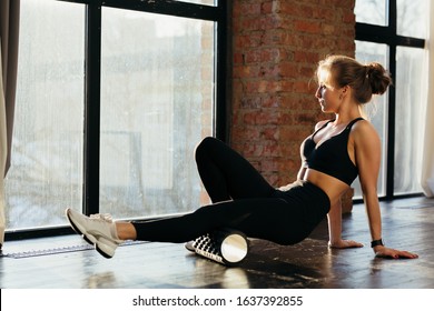 Fitness trainer girl in black leggings and a T-shirt in the training room flexes with a massage roll on the floor in the loft room