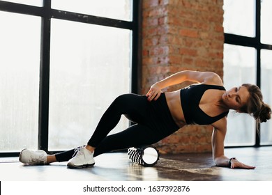 Fitness trainer girl in black leggings and a T-shirt in the training room flexes with a massage roll on the floor in the loft room