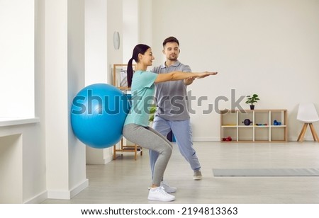Fitness trainer helping young female patient do wall squats with blue fit ball to get rid of backache and regain spinal health. Woman doing back exercise in physio room of modern clinic