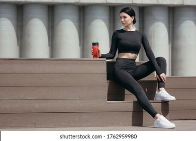 Fitness Trainer In A Sportwear Sits On The Bench And Rests After Jogging Holding A Red Drinking Bottle.