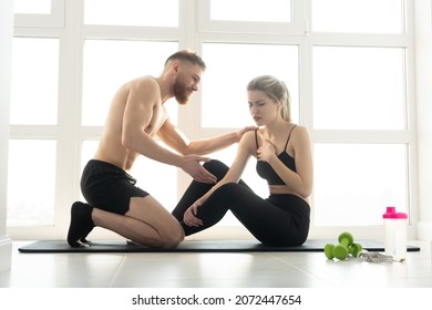 Fitness Trainer And Tired Sportswoman With Heart Pain After Training On Fitness Mat. Blonde Woman Wear Sportswear. Man With Naked Torso. Concept Of Sport Activity At Home. Sunny Spacious Apartment