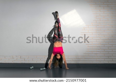 Fitness trainer works out in the gym. Young fit flexible woman wearing sportswear do handstand near the wall