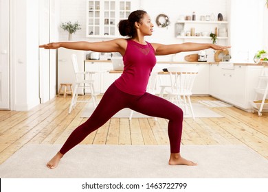 Fitness, training, health and wellness concept. Side view of athletic young Afro American housewife in stylish sportswear practicing yoga in the morning, doing warrior 2 pose on mat in kitchen