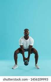 Fitness Training. Sports Man Doing Kettlebell Squat Workout On Blue Background. Black Male Athlete Exercising, Squatting With Kettle Bell At Studio