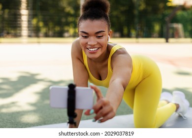 Fitness Vlog. Portrait Of Active Smiling Black Woman Filming Video For Her Internet Channel At Social Media, Using Smartphone On Tripod, Trainer In Earbuds Recording Tutorial On Sports Stadium