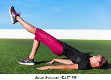 Fitness Woman Doing Bodyweight Glute Single Leg Floor Bridge Lift Exercices. Fit Asian Woman Exercising Glutes Muscles With One-legged Floor Bridge Butt Raise During Summer In Outdoor Gym On Grass.