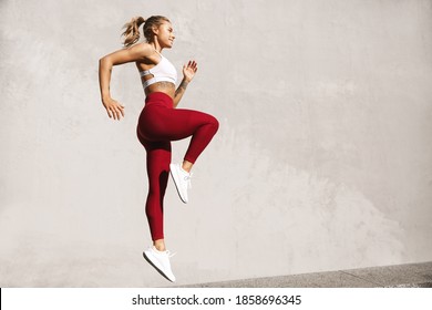 Fitness woman doing cardio interval training outdoors. Side view of athletic female model jumping by concrete wall, warm-up before workout and jogging. - Shutterstock ID 1858696345