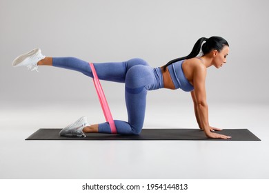 Fitness woman doing kickback exercise for glutes with resistance band on gray background. Athletic girl working out donkey kicks