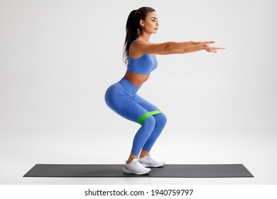 Fitness woman doing squats exercise for glute with resistance band on gray background. Athletic girl working out