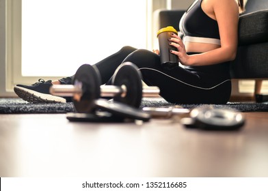 Fitness Woman Drinking Protein Or Recovery Drink After Workout And Weight Training. Working Out And Exercise At Home. Lady In Sportswear Holding Shaker. Dumbbell And Gym Equipment On The Floor.