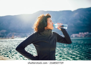 Fitness woman drinking water from bottle. Muscular young female taking a break from workout outside. - Powered by Shutterstock