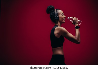 Fitness woman drinking water on red background. Smiling african female in sportswear and headphones drinking water after exercising.