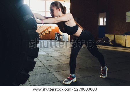 Fitness woman flipping wheel tire in gym. Fit female athlete working out with a huge tire. Back view. Sportswoman doing an strength exercise training.