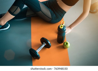 Fitness Woman With Healthy Workout Equipment On Gym Floor. Exercise And Body Build Up Concept. Beauty And Sport Theme. Relax And Rest Theme. Green Apple And Protein Shake Bottle And Dumbbell In Center