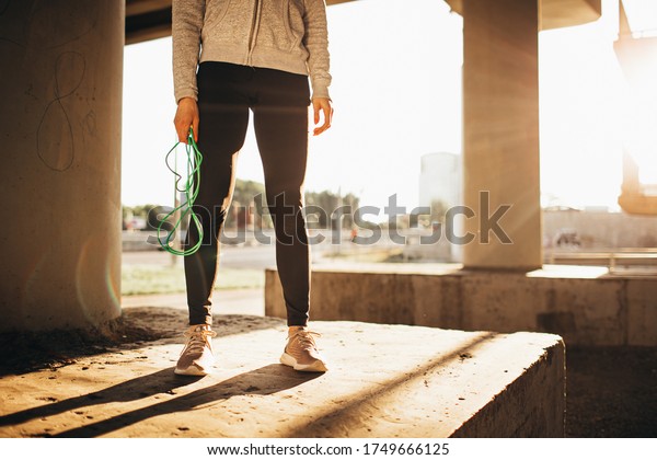 Fitness woman jump rope under a bridge at sunset\
preparitn to jump or take a rest after junping. Face is not\
visible. Outside exercises,\
cardio