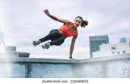 Fitness woman jumping on to the terrace from the rooftop fence with one hand on fence. Woman in fitness wear doing fitness training on rooftop. - Shutterstock ID 1256404051