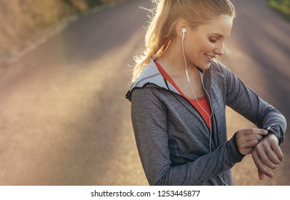 Fitness Woman Looking At Her Wrist Watch During Her Morning Fitness Run. Smiling Female Athlete Checking Time Standing On Road While Listening To Music On Earphones.