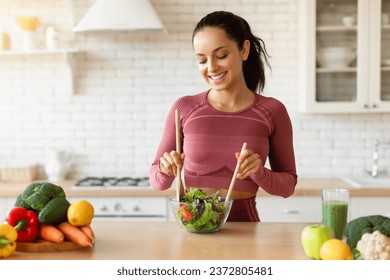 Fitness woman making a fresh salad in her kitchen at home, slimming on a healthy dieting routine, enjoying nutritious meal preparation with weight loss, wearing fitwear standing indoor