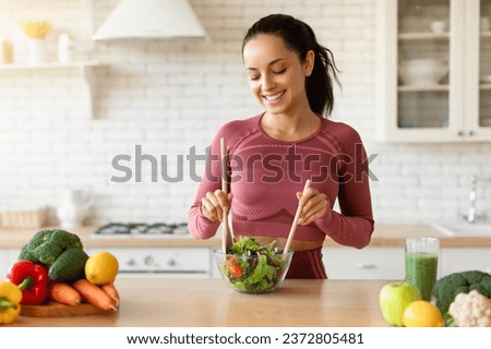 Fitness woman making a fresh salad in her kitchen at home, slimming on a healthy dieting routine, enjoying nutritious meal preparation with weight loss, wearing fitwear standing indoor