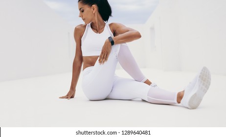 Fitness Woman Sitting On Floor And Twisting Her Back Sideways. Woman In White Fitness Clothes Holding One Leg Close And Turning To Her Side To Stretch Her Back.