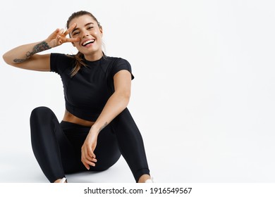 Fitness woman smiling and showing v-sign after workout. Healthy and fit female athlete resting, sitting on floor in sport clothes. Sportswoman enjoying productive exercising, white background. - Shutterstock ID 1916549567