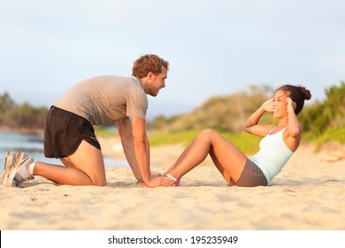 Fitness woman training situp crunches with personal trainer instructor. Young couple happy working out in sand on beach. Beautiful Asian female model and male fitness model holding her feet exercising - Powered by Shutterstock